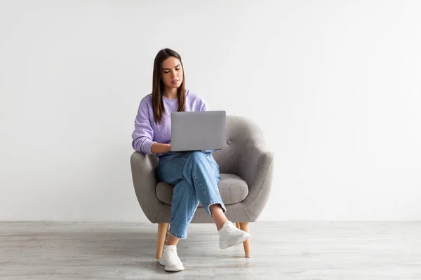 Sorridente giovane donna caucasica seduta su poltrona con computer portatile contro parete studio bianco, spazio copia — Foto Stock