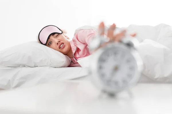 Funny sad tired millennial caucasian blonde lady in pink pajama and sleep mask reaches to alarm clock, lies on bed — Stock Photo, Image