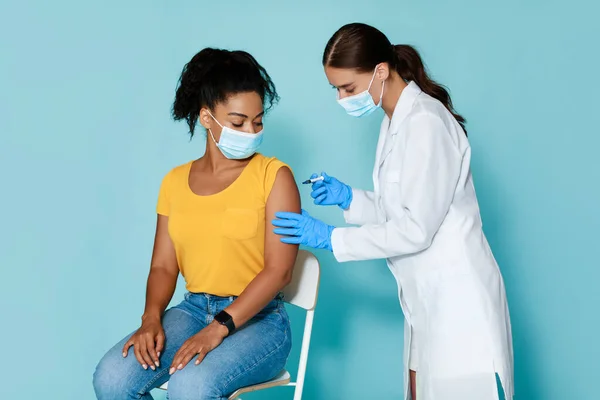 Médica mulher fazendo injeção de vacina contra o coronavírus para jovem senhora negra em máscara protetora, fundo estúdio azul — Fotografia de Stock