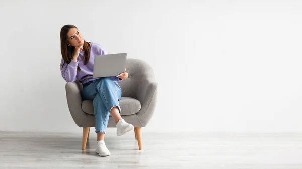 Mujer joven frustrada sentada en sillón con portátil, sintiéndose cansada después de un largo día de trabajo contra la pared blanca del estudio — Foto de Stock