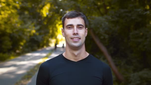 Außenporträt eines jungen motivierten Sportlers, der in die Kamera lächelt, posiert nach einem Spaziergang im grünen öffentlichen Park, Kamerafahrt — Stockvideo