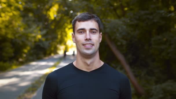 Ao ar livre retrato de jovem motivado desportivo cara sorrindo para a câmera, posando depois de andar no parque público verde, rastreamento tiro — Vídeo de Stock