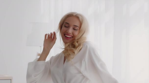 Mooie jonge blonde vrouw in witte zijden mantel dansen thuis — Stockvideo