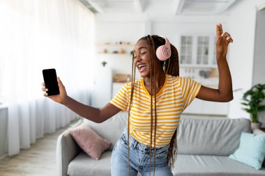 Neşeli Afro-Amerikan uzun saçlı kadın şarkı söylüyor ve dans ediyor, akıllı telefon kullanıyor.