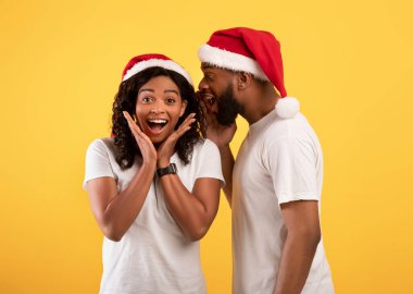 Siyahi adam sürpriz eşine yılbaşı sırrını fısıldıyor, kulağına büyü yapıyor, sarı arka planda Noel Baba şapkaları takıyor.