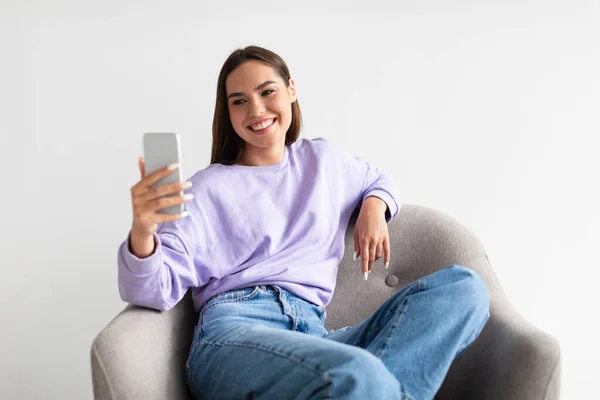 Mujer alegre usando teléfono inteligente, charlando en la web, trabajando o estudiando en línea, sentado en sillón sobre fondo blanco —  Fotos de Stock