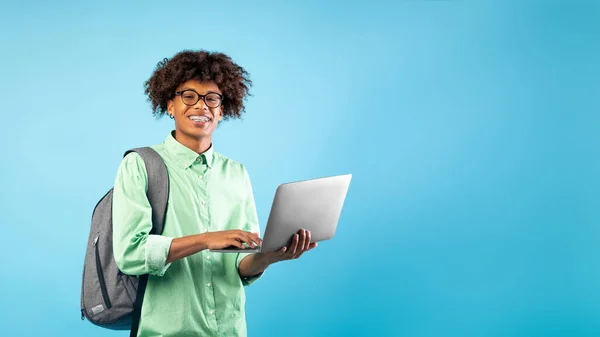 Koncepcja technologii i e-learningu. Podekscytowany czarny nastolatek facet z laptopem i plecakiem, niebieskie tło, puste miejsce — Zdjęcie stockowe