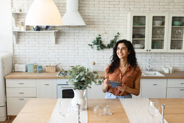 Glada unga brunett kvinna med hjälp av surfplatta dator, surfa internet eller sociala medier, arbetar online i köket — Stockfoto