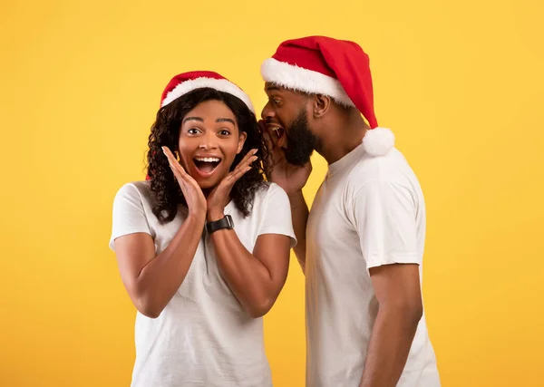 Svart man viskar jul hemlighet till överraskad fru, stavning till hennes öra, bär Santa hattar över gul bakgrund — Stockfoto
