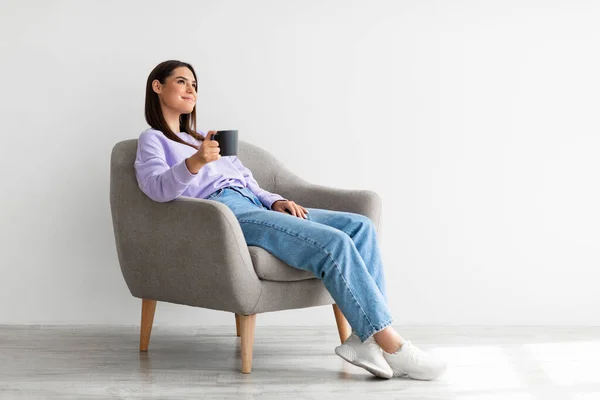 Mujer joven con taza de café caliente enfriamiento en sillón acogedor contra la pared del estudio blanco, espacio libre —  Fotos de Stock