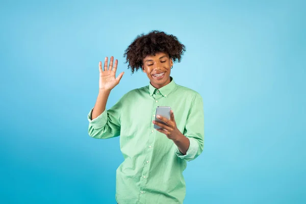 Feliz chico adolescente negro con teléfono inteligente haciendo videollamada, agitando la mano en la cámara del teléfono móvil, fondo azul — Foto de Stock