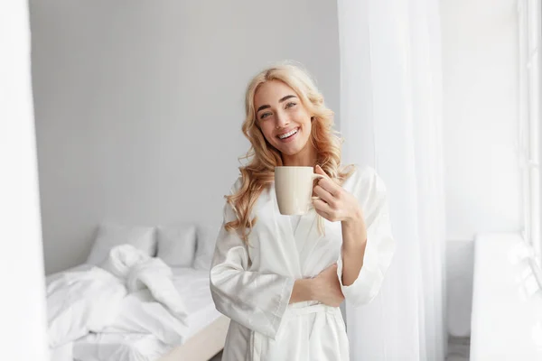 Ευτυχισμένη χιλιετή όμορφη καυκάσια γυναίκα με μπουρνούζι απολαμβάνει καλημέρα και πίνει νόστιμο φρέσκο καφέ — Φωτογραφία Αρχείου