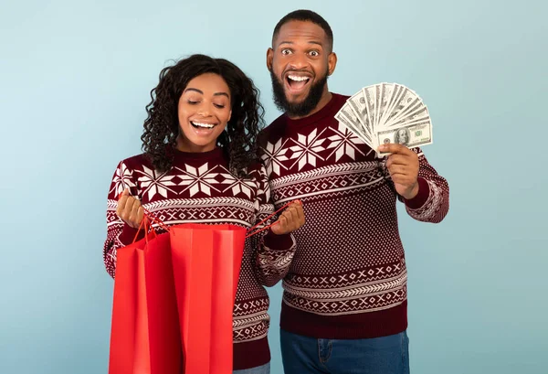 Kerstinkopen. Gelukkig Afrikaans Amerikaans paar in kerst truien met boodschappentassen en een hoop geld — Stockfoto