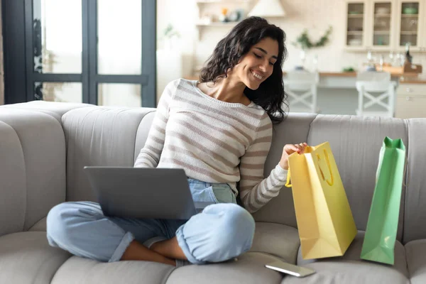 Joven mujer alegre sentada en el sofá con el ordenador portátil, mirando dentro de bolsas de compras, ordenar productos en línea desde casa — Foto de Stock
