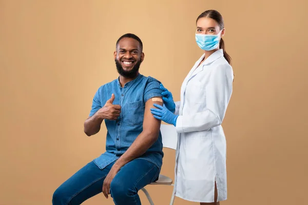 Medico donna in maschera e ragazzo di colore che mostra pollice in su dopo l'iniezione del vaccino, raccomandando l'immunizzazione contro covid — Foto Stock