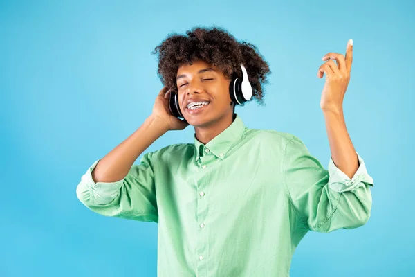 Joyeux noir adolescent gars écouter de la musique dans les écouteurs, fermer les yeux et se détendre sur fond bleu studio — Photo
