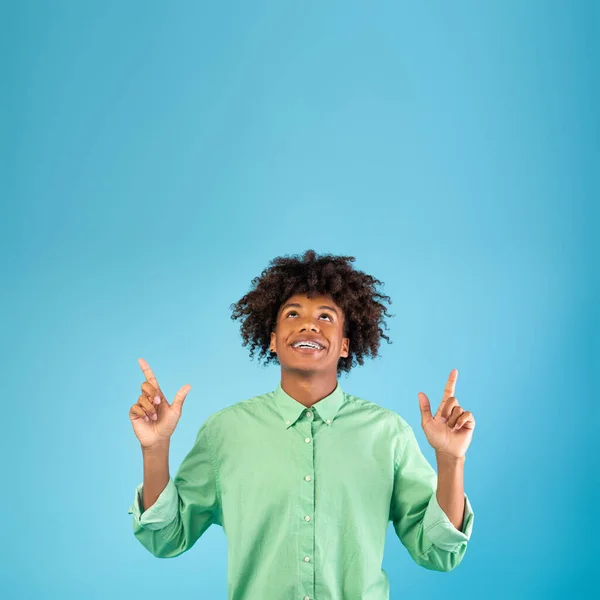 ¡Mira esto! Chico negro feliz señalando los dedos hacia arriba en el espacio vacío por encima de su cabeza, posando sobre el fondo azul, cosecha —  Fotos de Stock