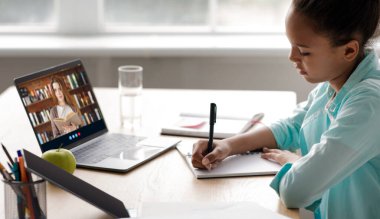 Genç Avrupalı kadın öğretmen dizüstü bilgisayar ekranında. Genç Afro-Amerikalı kız evde online ders çalışıyor.