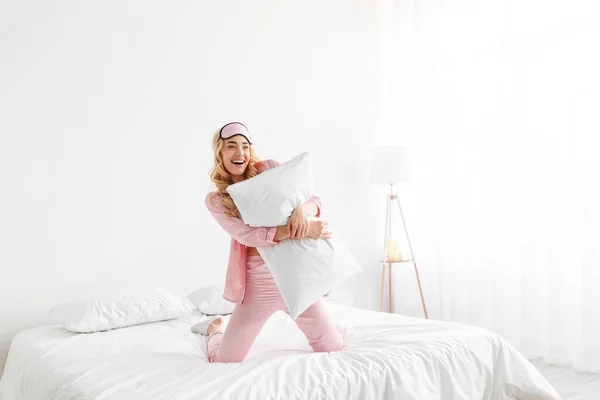 Rolig glad tusenårig söt kaukasiska blond kvinna i rosa pyjamas, sömnmask, kram kudde, ha kul på komfortsäng — Stockfoto