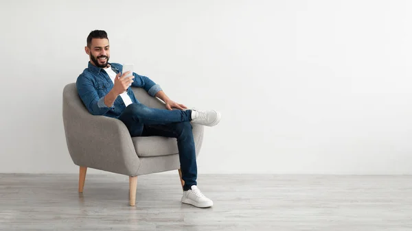 Neşeli genç Arap adam akıllı telefonuyla online iş görüşmesi yapıyor, beyaz duvarın yanındaki rahat koltukta oturuyor. — Stok fotoğraf