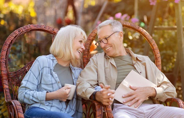 Romántica pareja de ancianos sentados en el jardín en sillas de mimbre, mirando a eah otros con amor y disfrutando de cálidos días de otoño —  Fotos de Stock
