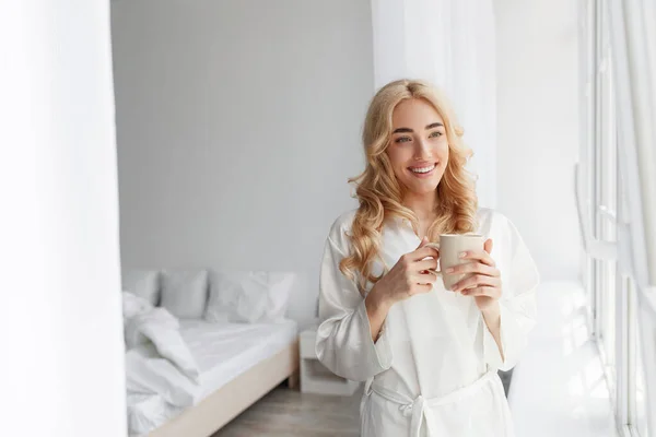 Vrolijk jong schattig europees blond vrouw in badjas genieten van goedemorgen, rust, stilte en drink heerlijke verse koffie — Stockfoto