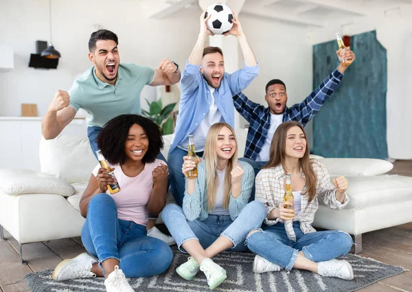 Amigos emocionales diversos viendo fútbol en la televisión, bebiendo cerveza, sosteniendo la pelota y gritando, celebrando la victoria en casa — Foto de Stock