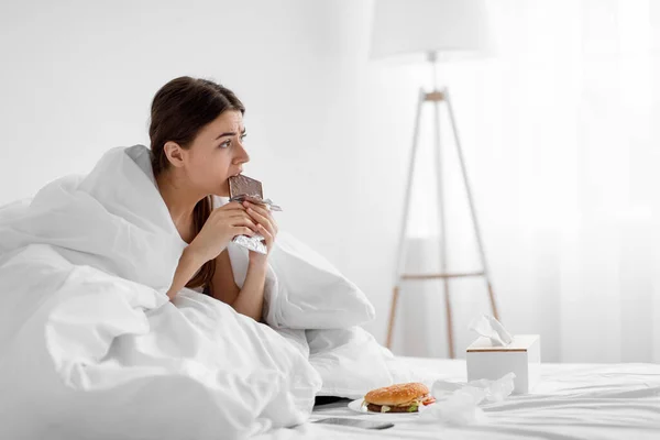 Rozczarowana zdenerwowana tysiącletnia kobieta jedząca czekoladę w łóżku z fast foodem w domu, cierpiąca z powodu przejedzenia — Zdjęcie stockowe