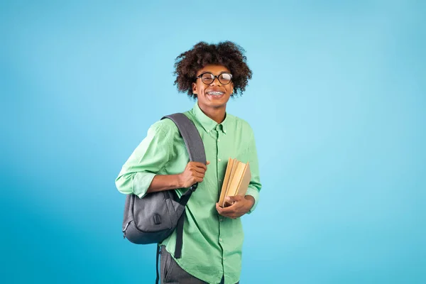 Felice nero ragazzo studente con zaino e libri sorridente a fotocamera su blu studio sfondo — Foto Stock