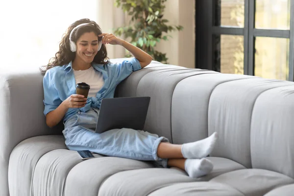 Felice giovane donna caucasica utilizzando laptop e cuffie, avendo videochiamata online, bere caffè a casa — Foto Stock