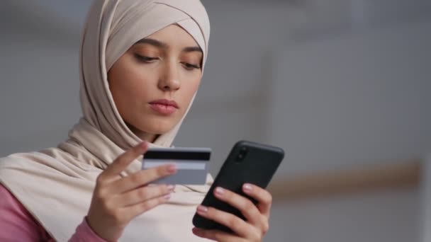 Молодая мусульманка в платке совершает онлайн-транзакцию на смартфоне дома, замедленная съемка, пустое пространство — стоковое видео