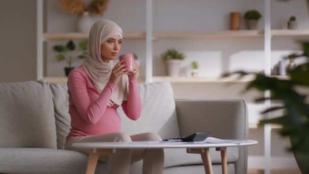 Ung gravid Mellanösternkvinna i hijab koppla av hemma, dricka hälsosamt te och smeka hennes stora mage — Stockvideo