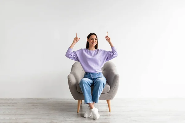 Mujer sonriente sentada en sillón, apuntando hacia arriba con ambas manos, ofreciendo espacio vacío sobre su cabeza, maqueta —  Fotos de Stock