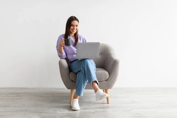 Позитивная белая женщина, работающая онлайн, сидящая в кресле с кофе и использующая ноутбук против белой стены студии — стоковое фото