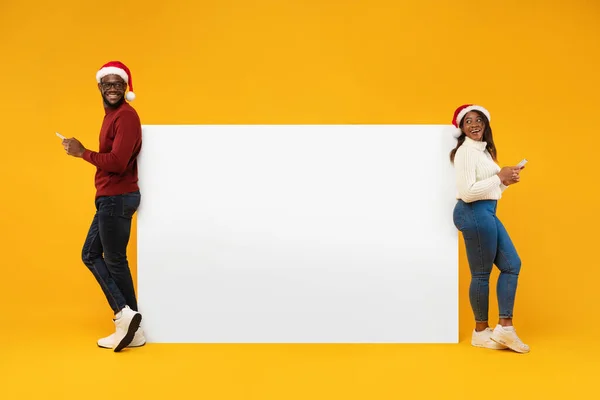 Pareja negra usando teléfonos inteligentes cerca de cartel para la Navidad Anuncio, Estudio — Foto de Stock