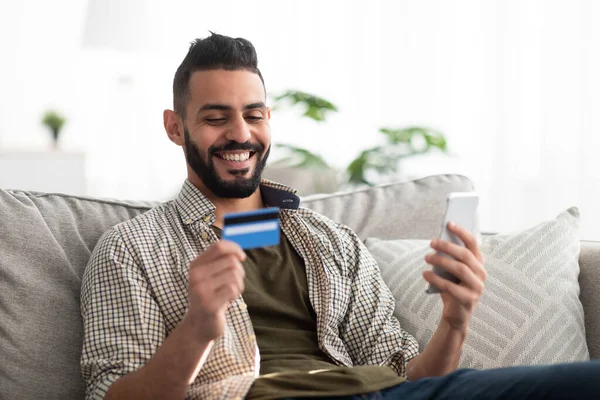 Joven árabe positivo usando tarjeta de crédito y teléfono inteligente para compras en línea, utilizando el servicio de banca remota en casa —  Fotos de Stock