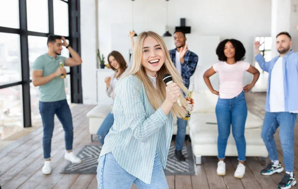 Jovem loira alegre com garrafa de cerveja e microfone cantando karaoke com amigos diversos em casa — Fotografia de Stock