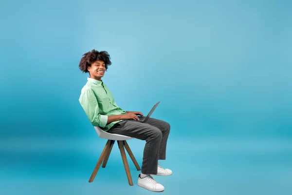 Concepto de aprendizaje electrónico. negro chico estudiante usando laptop ordenador, sentado en silla sobre azul estudio fondo, vacío espacio — Foto de Stock