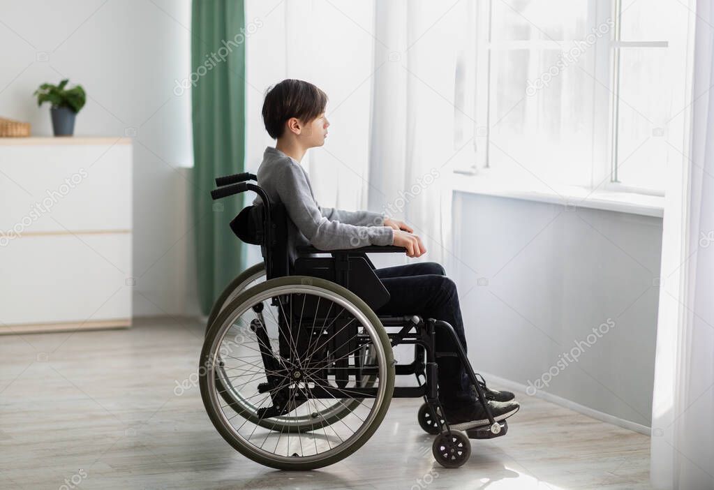 Unhappy impaired teenage boy sitting in wheelchair, looking out window, feeling desperate and alone at home