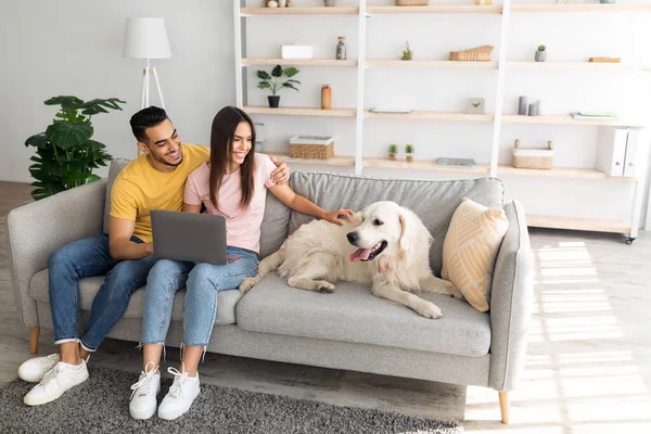 Alegre joven pareja multirracial sentado en el sofá con el ordenador portátil, acariciando a su perro en casa, espacio para copiar — Foto de Stock