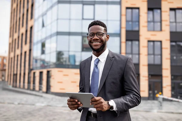 Nouvelles d'affaires. Heureux homme d'affaires noir utilisant une tablette numérique marchant à l'extérieur près d'un immeuble de bureaux moderne, espace de copie — Photo