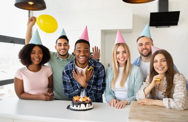Загадай желание. Позитивные разнообразные молодые друзья в шляпах для вечеринок празднуют вместе, позируя с тортом на день рождения дома — стоковое фото