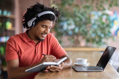 Genç siyahi adam kafede dizüstü bilgisayarla çalışırken not alıyor.