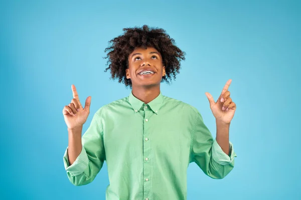Positivo afro-americano apontando os dedos acima de sua cabeça, posando sobre fundo azul — Fotografia de Stock
