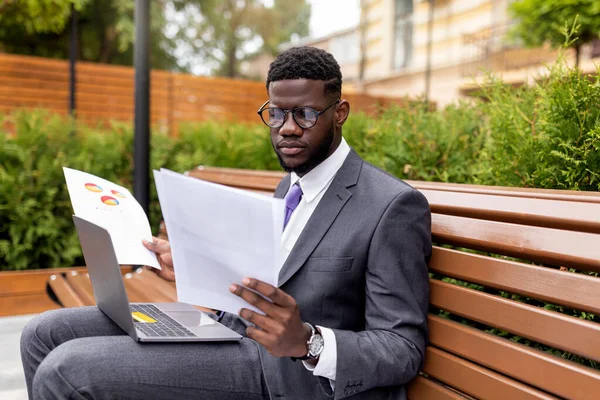 Συγκεντρωμένος Αφροαμερικανός επιχειρηματίας που κάθεται δίπλα στο γραφείο με laptop και έγγραφα ανάγνωσης, ελεύθερος χώρος — Φωτογραφία Αρχείου