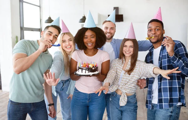 Retrato de diversos amigos jóvenes con sombreros festivos y sopladores de fiestas celebrando pastel de cumpleaños, mirando a la cámara en el interior —  Fotos de Stock