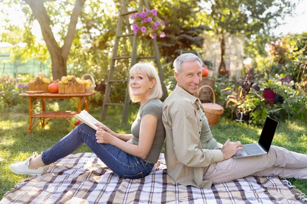 Щаслива старша пара відпочиває на пікніку ковдру в саду, чоловік використовує ноутбук, жінка читає книгу — стокове фото