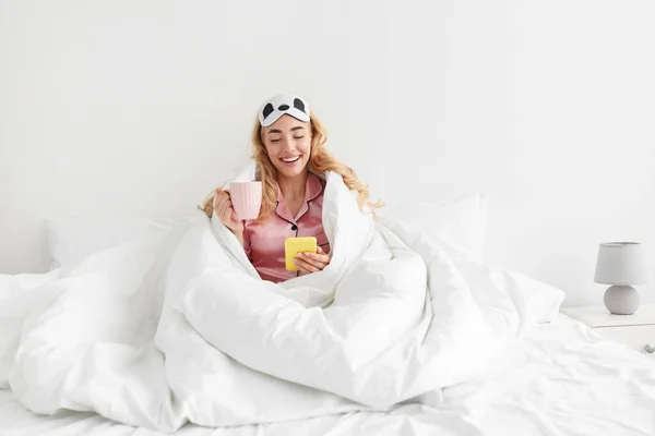Divertida mujer rubia caucásica milenaria en pijama rosa y máscara del sueño disfruta del café y lee el mensaje en el teléfono inteligente —  Fotos de Stock