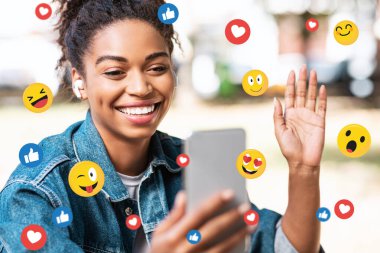 Kulaklıklı mutlu siyah milenyum kadını görüntülü konuşma yapıyor, emoji ile çifte çekim yapıyor.