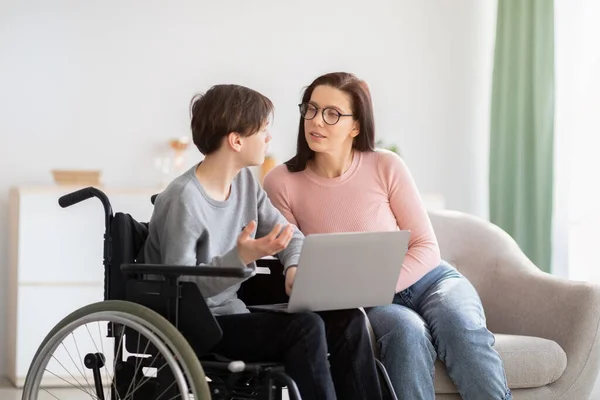 Web-based education. Cheerful disabled teen boy in wheelchair, making home assignment with his mom, using laptop indoors